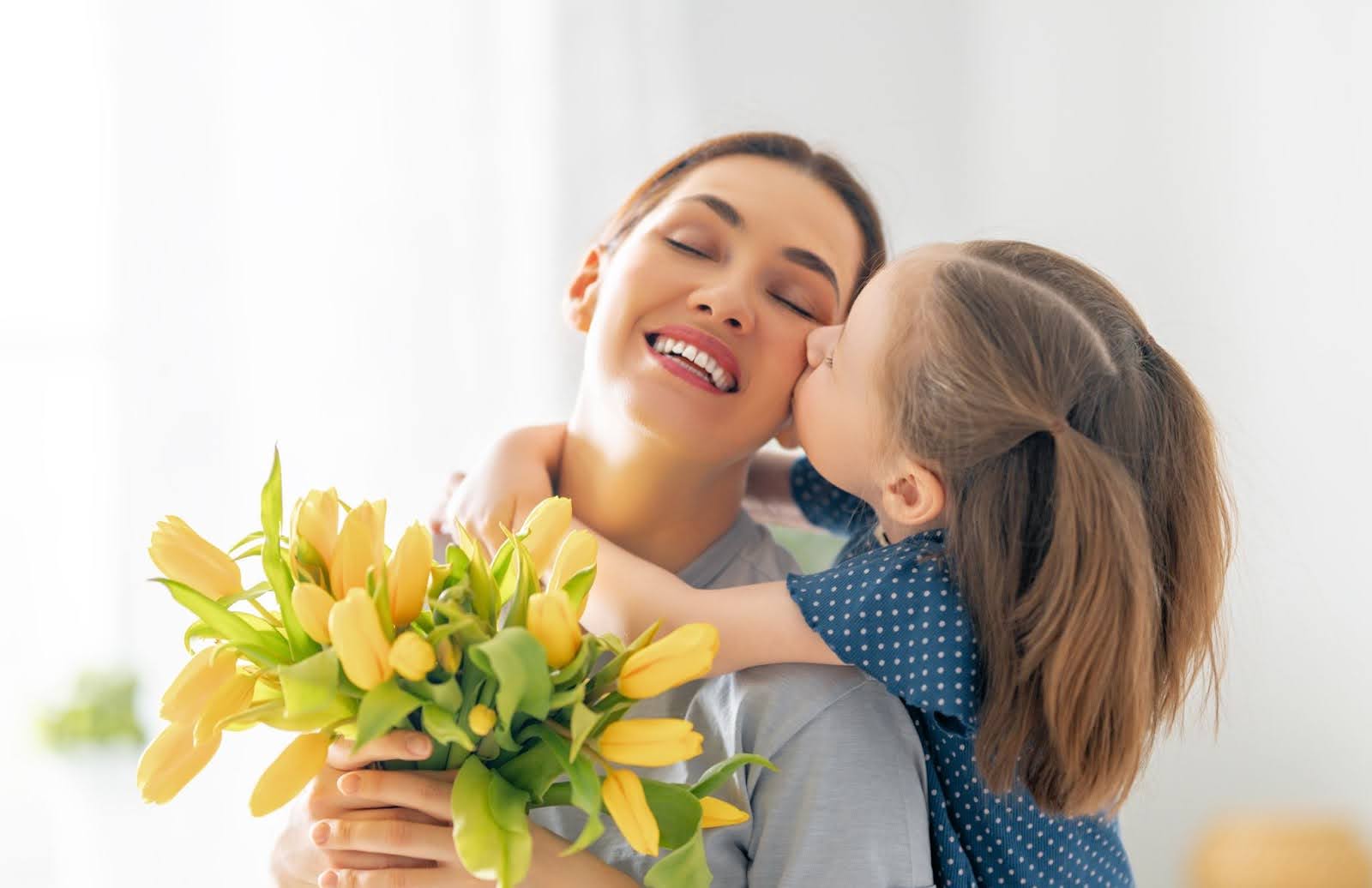 A child hugs and kisses her mother while presenting with a beautiful Mother's Day flower bouquet.